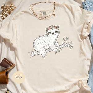 Può includere: T-shirt color avorio con un bradipo dei cartoni animati con una corona di fiori, appollaiato su un ramo. Il bradipo ha un'espressione scontrosa. La maglietta ha la scritta "Comfort Colors".