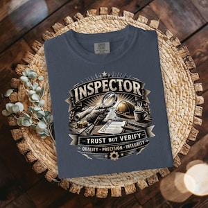 May include: A navy blue t-shirt with a graphic design featuring the word "INSPECTOR" above a magnifying glass, hard hat, and tools. The text "TRUST BUT VERIFY" and "QUALITY PRECISION INTEGRITY" are also included.