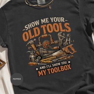 Könnte beinhalten: Dunkelgraues T-Shirt mit einem Vintage-Design, das verschiedene Werkzeuge wie Hammer und Säge zeigt. Der Text lautet "SHOW ME YOUR OLD TOOLS AND I'LL SHOW YOU MY TOOLBOX" in Orange und Weiß.