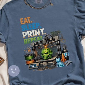 Può includere: T-shirt blu jeans con la scritta "EAT. SLEEP. PRINT. REPEAT." È presente una grafica di una stampante 3D, insieme a una testa di robot verde, un gatto blu e un razzo. La maglietta ha anche una bobina di filamento verde.
