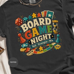 May include: A charcoal gray t-shirt featuring a colorful graphic with the text "BOARD GAME NIGHT" and "Roll • Strategize • Win". The design includes illustrations of dice, game pieces, an hourglass, playing cards, and a circular game board path.