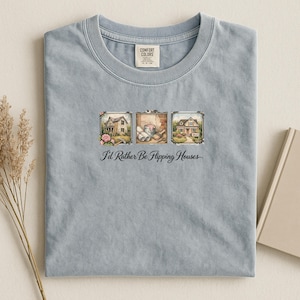 May include: A light blue Comfort Colors t-shirt featuring a graphic with three framed images of houses. The text "I'd Rather Be Flipping Houses" is printed below the images. The shirt is folded on a light surface.
