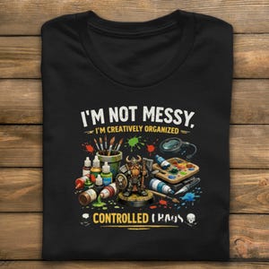 May include: Black t-shirt with the text "I'M NOT MESSY, I'M CREATIVELY ORGANIZED = CONTROLLED CHAOS." The shirt features a colorful graphic of paint bottles, brushes, a miniature figure, and a magnifying glass.