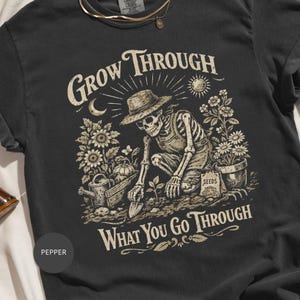 May include: Dark grey t-shirt with a skeleton gardener design and the text "Grow Through What You Go Through." The illustration includes sunflowers, a watering can, and a seed packet. A comfortable, casual wear.