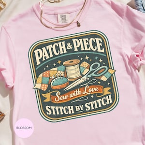 May include: A pale pink t-shirt with a sewing-themed graphic. The design includes the words "PATCH & PIECE", "STITCH BY STITCH", and "Sew with Love". The graphic also features a spool of thread, scissors, a pincushion, and a patchwork design.