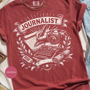 May include: A crimson short-sleeve t-shirt featuring a white graphic print of a hand holding a pen over a notepad, a camera, a typewriter, a microphone, and a rolled newspaper. The word JOURNALIST is printed in a banner above the illustrations.