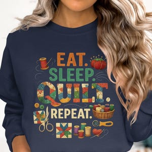 May include: Navy blue sweatshirt with the text "EAT. SLEEP. QUILT. REPEAT." in colorful letters. The design includes illustrations of sewing notions such as spools of thread, scissors, and a tomato pin cushion. A cozy garment for quilters.