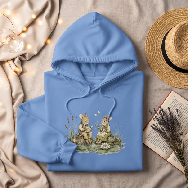 Blue hoodie with a watercolor illustration of two rabbits by a pond.