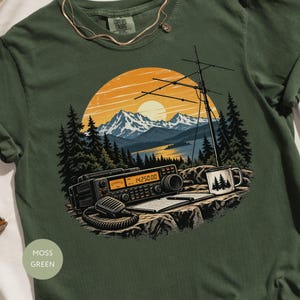 May include: A moss green t-shirt featuring a graphic of a radio transceiver, a microphone, a notebook, and a mug with a pine tree print. The background shows a mountain landscape with a large orange sun, pine trees, and a tall antenna tower.