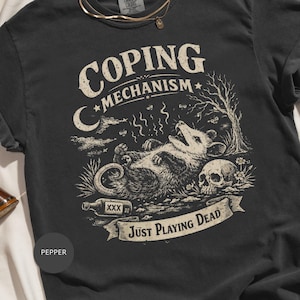 Puede incluir: Camiseta gris oscuro con un gráfico desgastado que presenta una zarigüeya tumbada, una calavera y el texto "Just Playing Dead". La camiseta también tiene las palabras "Coping Mechanism".