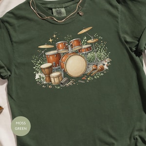 Può includere: T-shirt verde muschio con un'illustrazione di batteria. Il design include tamburi marroni, piatti e maracas. La maglietta ha un girocollo e maniche corte. Il testo "Comfort Colors" è visibile sul colletto.
