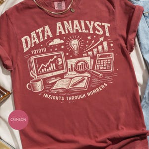 May include: A crimson short-sleeve t-shirt featuring a cream-colored graphic print. The design includes the text "DATA ANALYST" and "INSIGHTS THROUGH NUMBERS" alongside illustrations of a computer monitor, a lightbulb, a calculator, and a book.