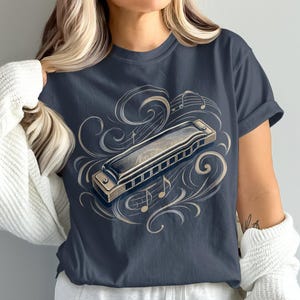 May include: A dark gray short-sleeve t-shirt featuring a central graphic of a metallic harmonica surrounded by light-colored swirling filigree patterns and musical notes. The design is centered on the chest of the garment.