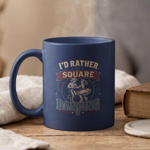 May include: A navy blue ceramic mug featuring a graphic print of a couple square dancing. The text reads I'D RATHER BE SQUARE DANCING in white and red lettering. The design includes stars and decorative flourishes in a vintage-inspired style.