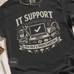 May include: A charcoal gray t-shirt featuring a cream-colored graphic print. The design includes a laptop with a checkmark, a computer tower, a wrench, a screwdriver, headphones, a coffee mug, and a mouse, with the text IT SUPPORT TECH HELP & TROUBLESHOOTING.