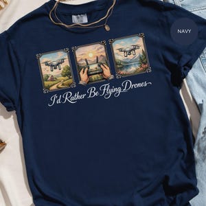 Puede incluir: Camiseta azul marino con un gráfico que presenta tres escenas de drones enmarcadas. Las escenas representan un dron sobre una granja, manos sosteniendo un controlador al atardecer y un dron sobre un lago. El texto "I'd Rather Be Flying Drones" está debajo.