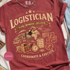 Könnte beinhalten: Ein karmesinrotes Kurzarm-T-Shirt mit einer verwaschenen goldenen Grafik mit dem Text LOGISTICIAN, PLAN, MANAGE, DELIVER und COORDINATE & EXECUTE. Das Design zeigt einen Sattelzug, ein Flugzeug, einen Gabelstapler, eine Weltkarte und einen Kompass.