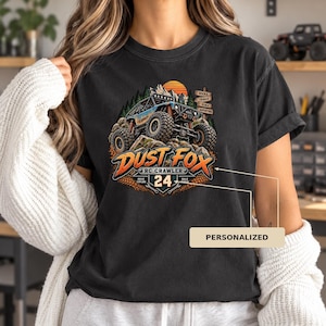 May include: A black t-shirt featuring a graphic of a blue RC crawler vehicle on rocky terrain with a sunset, pine trees, and a signpost. The text reads DUST FOX, RC CRAWLER, 24, DRIVE, EXPLORE, BUILD, REPEAT, ROCKS, TRAILS, SCALE, ADVENTURE.
