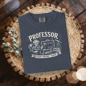 May include: A navy blue t-shirt with a graphic featuring the word "PROFESSOR" above an illustration of a professor, a chalkboard with the equation E=mc², and the text "LEARN • INSPIRE • TEACH".