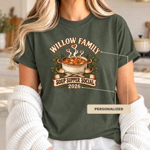 Op de afbeelding: Een olijfgroen t-shirt met een afbeelding van een soeppan met twee houten lepels, omringd door groen en stoomhartjes. De tekst luidt WILLOW FAMILY SOUP SUPPER SOCIAL 2026. Een beige vakje met het woord PERSONALIZED is zichtbaar.