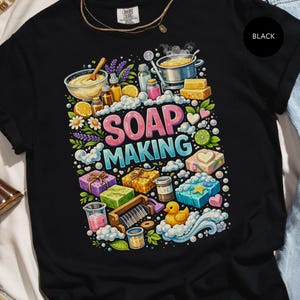 Può includere: T-shirt nero con un design grafico colorato. Il design presenta le parole "SOAP MAKING" in rosa e blu, circondate da ingredienti e strumenti per la produzione di sapone, tra cui oli essenziali, una pentola e una paperella di gomma.