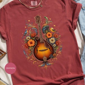 May include: Crimson-colored t-shirt featuring a detailed illustration of a mandolin surrounded by flowers and birds. The mandolin has a brown and yellow finish. The design includes orange, blue, and yellow flowers, with a few birds. The shirt is made of a soft material.