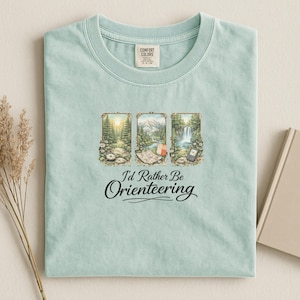 May include: A light blue t-shirt with the text "I'd Rather Be Orienteering" in a cursive font. The shirt features three framed illustrations of outdoor scenes, including a compass, map, and waterfall. The shirt is made of a soft material.