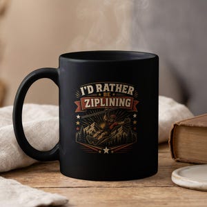 May include: A matte black ceramic mug featuring a graphic illustration of a person ziplining over a mountain forest. The text "I'D RATHER BE ZIPLINING" is printed in a bold, vintage-style font above the illustration, accented by stars and banners.