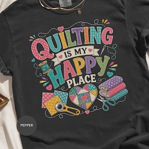 May include: A charcoal gray t-shirt featuring a colorful graphic with the text QUILTING IS MY HAPPY PLACE. The design includes illustrations of fabric swatches, a rotary cutter, scissors, thread spools, and a heart-shaped quilt block pattern.