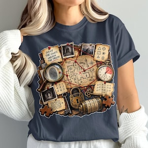 Puede incluir: Una camiseta gris oscuro con un estampado gráfico de un tablero de misterio con temática de detectives. El diseño incluye un mapa, una lupa con una huella dactilar, un cronómetro, un candado, una llave, un disco de cifrado y notas con el texto Solve the clues unlock the truth.
