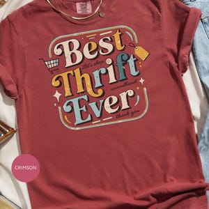May include: Crimson-colored t-shirt with the text "Best Thrift Ever" in a retro design. The graphic includes a shopping cart, a price tag, and the words "Let's shop secondhand, thank you."