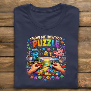 以下が含まれることがあります： 「SHOW ME HOW YOU PUZZLE」の文字が入った、カラフルなグラフィックが特徴のネイビーブルーのTシャツ。デザインには、漫画のキャラクター、風景、パズルを組み立てる手などが含まれています。Tシャツは木製の表面に折りたたまれています。