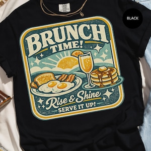 Könnte beinhalten: Schwarzes T-Shirt mit einem Retro-Brunch-Design. Das Design zeigt die Worte "Brunch Time!" über Illustrationen von Frühstücksgerichten, darunter Pfannkuchen, Eier und Toast. Der Satz "Rise & Shine Serve It Up!" ist ebenfalls enthalten.