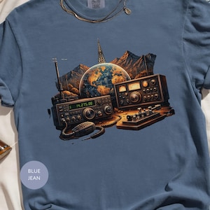 May include: Blue jean colored t-shirt featuring a graphic of vintage radio equipment, a globe, and mountains. The graphic includes the text "14.275.00". The shirt has a crew neck and short sleeves.
