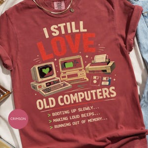 May include: A crimson t-shirt featuring a graphic of vintage computer equipment, including a monitor with a heart icon, a keyboard, and a printer. The text reads: I STILL LOVE OLD COMPUTERS, BOOTING UP SLOWLY, MAKING LOUD BEEPS, RUNNING OUT OF MEMORY.