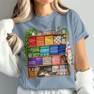 Puede incluir: Una camiseta azul claro con un estampado gráfico de una estantería de madera llena de pilas de telas plegadas de colores, plantas en macetas, artículos de costura y un gato atigrado durmiendo en el estante inferior, con colores del arcoíris.