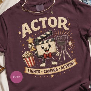 May include: A berry-colored t-shirt featuring a graphic of a cartoon film clapperboard character wearing a red bow tie, holding a gold star trophy, standing next to a movie camera and popcorn bucket, with the text ACTOR and LIGHTS CAMERA ACTION.