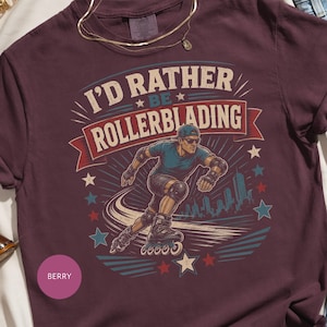 Peut inclure: Un t-shirt couleur baie foncée avec un motif représentant une personne faisant du roller. Le texte indique I'D RATHER BE ROLLERBLADING. Le design inclut une silhouette urbaine, des étoiles et une bannière rouge. Col rond et manches courtes.