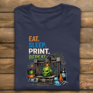 May include: Navy blue t-shirt with the words "EAT. SLEEP. PRINT. REPEAT." in colorful text. The graphic features a 3D printer with a green helmet, a blue cat, and other 3D printing related items.