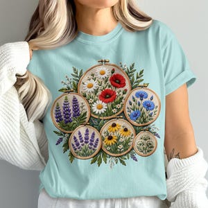 May include: A light blue t-shirt featuring a graphic print of six circular embroidery hoops. Each hoop displays a different floral arrangement, including red poppies, white daisies, purple lupines, blue cornflowers, yellow coneflowers, and white yarrow.