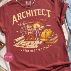 Puede incluir: Una camiseta de manga corta color carmesí con un estampado gráfico que incluye el texto ARCHITECT y DESIGNING THE FUTURE. La ilustración muestra una grúa de construcción, un edificio, un compás, un casco amarillo, libros y planos.