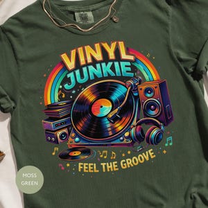 Puede incluir: Camiseta verde musgo con un gráfico colorido de un tocadiscos, discos, altavoces y auriculares. El texto "VINYL JUNKIE" está sobre un arcoíris, con "FEEL THE GROOVE" debajo. Etiqueta Comfort Colors visible.