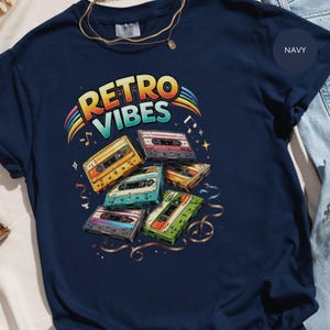 Può includere: T-shirt blu navy con grafica colorata "Retro Vibes". Il design presenta un arcobaleno, note musicali e diverse musicassette in vari colori. Una collana d'oro è visibile vicino allo scollo.