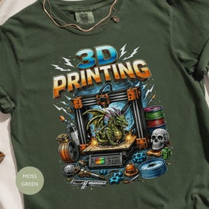 Op de afbeelding: Mosgroen t-shirt met een kleurrijke afbeelding van een 3D-printer, een draak en de tekst "3D PRINTING". Het ontwerp bevat een schedel, spoelen filament en diverse gereedschappen. Het shirt is gemaakt van zacht materiaal.
