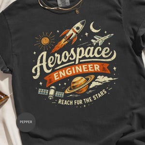 May include: Pepper-colored t-shirt with a vintage-style graphic. The design features a rocket, sun, planet, and the words "Aerospace Engineer" and "Reach for the Stars." The graphic is in shades of orange, white, and beige.