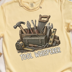 May include: A light yellow t-shirt with a graphic of various tools, including a hammer, saw, and wrench, inside a wooden box and a toolbox. The text "TOOL WHISPERER" is printed below the tools.