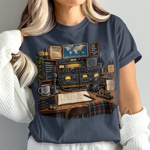 May include: A dark grey t-shirt featuring a graphic illustration of an amateur radio station setup. The design includes radio equipment, a world map, a logbook, a microphone, headphones, and various text elements like Amateur Radio and Radio Connects the World.