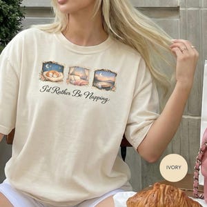 May include: Ivory-colored t-shirt with the text "I'd Rather Be Napping." The shirt features three framed illustrations: a sleeping cat, a bed, and a sunset. The shirt is made of a soft material.