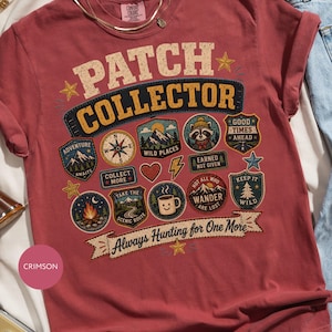 May include: A crimson short-sleeve t-shirt featuring the text PATCH COLLECTOR and Always Hunting for One More. The graphic includes various colorful patches with illustrations of mountains, a compass, a campfire, a raccoon, and a coffee mug.