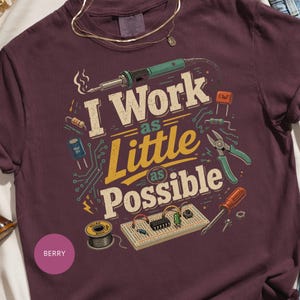 以下が含まれることがあります： ベリー色のTシャツで、はんだごて、ワイヤーカッター、ドライバー、ブレッドボードなどの電子部品のグラフィックが描かれています。テキストは「I Work as Little as Possible」とあり、回路基板のイラストで囲まれています。
